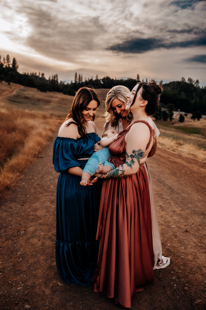 Family Photographer, a mother holds her baby as two other woman, sisters admire them on a country road. They all wear dresses.