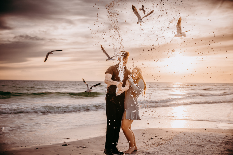 Family Photography, a man and woman stand at the beach popping a bottle of champagne, birds fly around them